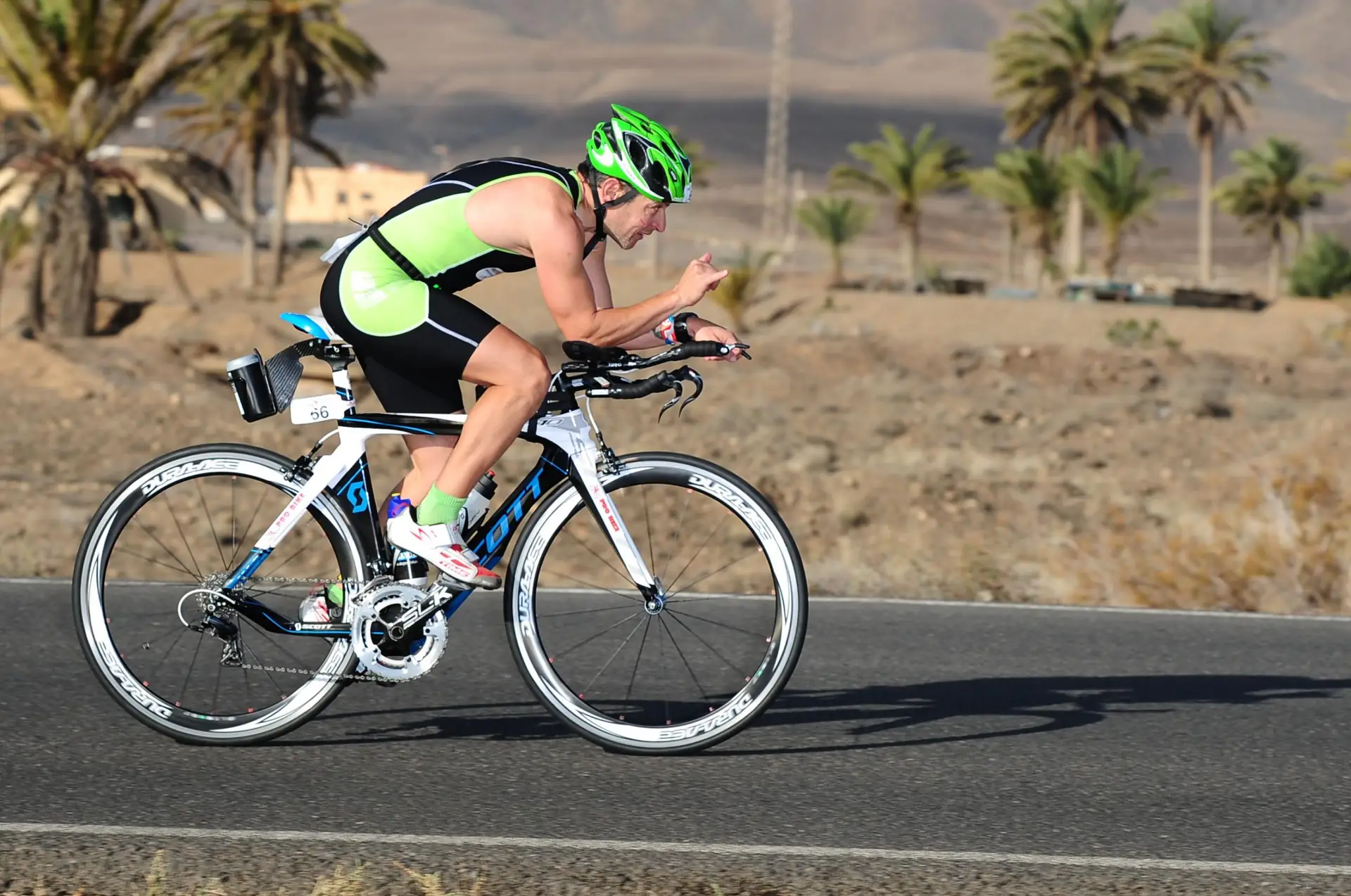 Triatleta con casco verde en bicicleta de contrarreloj por una carretera de Lanzarote, con palmeras al fondo